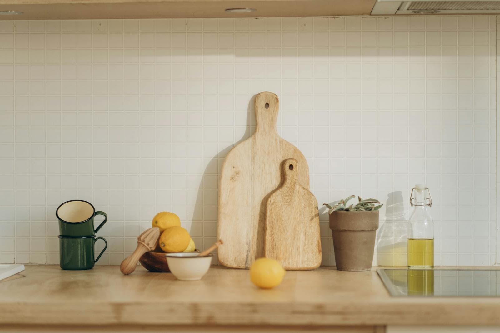 Wooden cutting boards, enamel mugs, a ceramic citrus reamer with lemons, and an olive oil bottle arranged on a sunlit kitchen counter