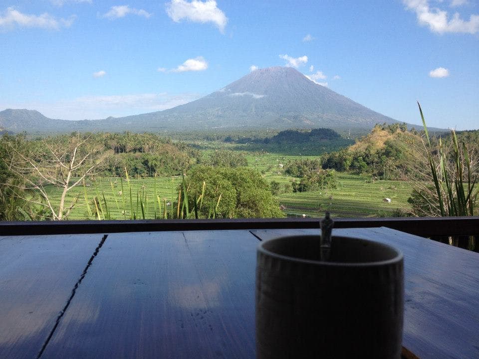A steaming mug sits on a wooden table overlooking a lush green valley with a majestic volcanic mountain in the distance under a partly cloudy sky.